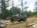 1987 Suzuki Samurai - A blue Jeep Cherokee XJ tackles a challenging rocky obstacle on a muddy off-road trail, with another vehicle visible in the background.