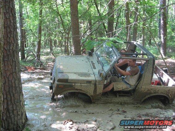 Muddy 1987 Suzuki Samurai stuck in a forest puddle, 3q-front view