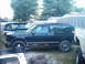 1994 GMC Jimmy - A 1994 GMC Jimmy is seen from the side in a yard, with visible rust on its lower body panels and other vehicles nearby.