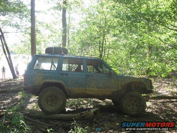 Teal Jeep Cherokee XJ with spare tire on roof driving over logs in muddy woods