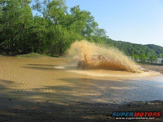 Off-road vehicle driving through a large mud puddle, kicking up a lot of water