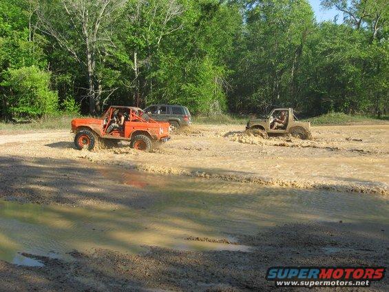 Orange off-road vehicle and tan off-road vehicle driving through mud, with a dark SUV behind