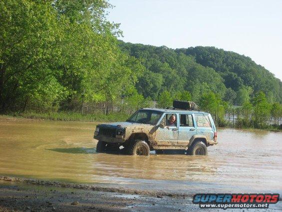 Light blue Jeep Cherokee XJ with mud on it driving through shallow water, spare tire on roof rack