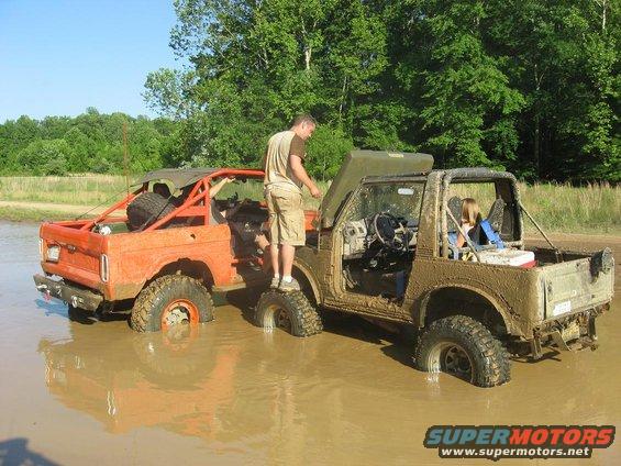 Two off-road vehicles stuck in mud. One is orange with a roll cage, the other is tan with its hood up