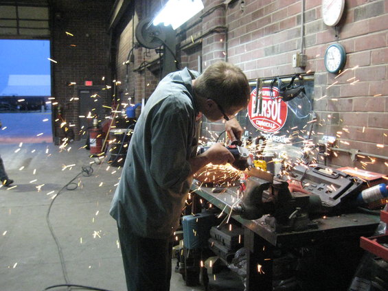Man in workshop using angle grinder, sparks flying. "ELR SOL CHEMICALS" sign visible