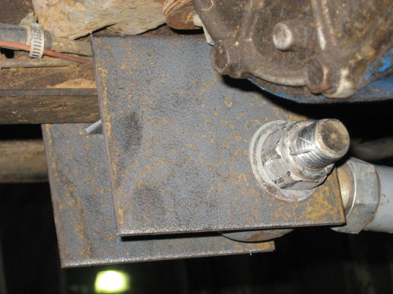 Undercarriage of a vehicle showing a rusty metal bracket bolted to a frame, with part of the drivetrain visible