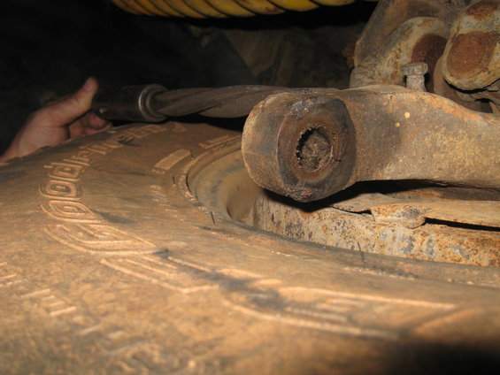 Close-up of a vehicle's rusty suspension linkage and a tire. A hand holds a tool on a cable