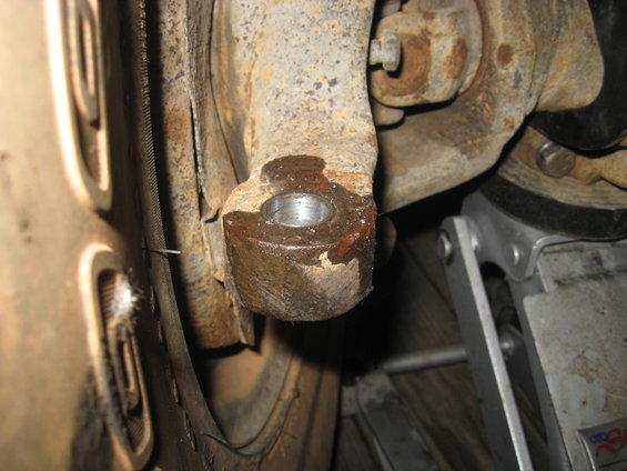 Close-up of a heavily rusted suspension component, possibly a tie rod end, with a silver hydraulic jack visible to the right