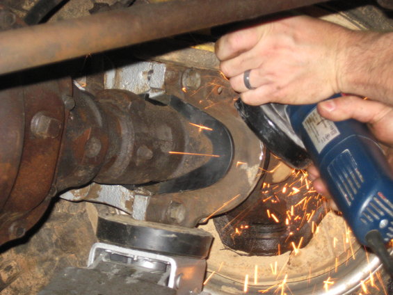 Hands with a ring using a blue angle grinder to cut metal near a rusty drivetrain with sparks flying
