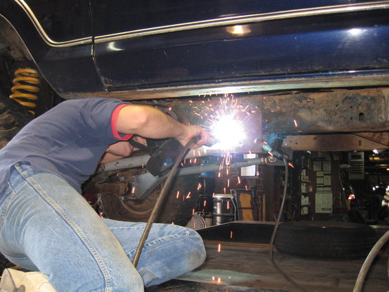 Person welding the frame of a dark blue vehicle, with a yellow spring visible