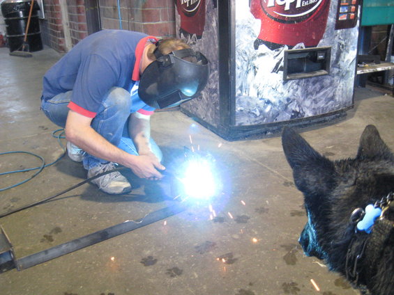 Man in blue shirt and jeans welding metal bar, dog in foreground, vending machine behind