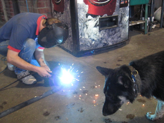 Man in welding mask and jeans welds metal bar, dog watches nearby