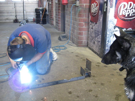 Man in welding mask working on metal piece on floor, dog watches nearby, Dr Pepper machine in background