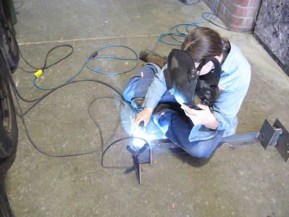 Person in jeans and a light blue shirt welding metal parts on a concrete floor. Welding mask is up, showing their face. Welding equipment and cords are visible