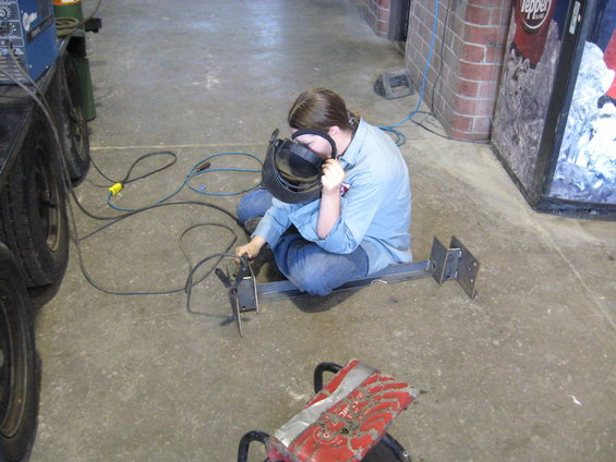 Person in blue shirt and jeans welding metal brackets while sitting on the floor