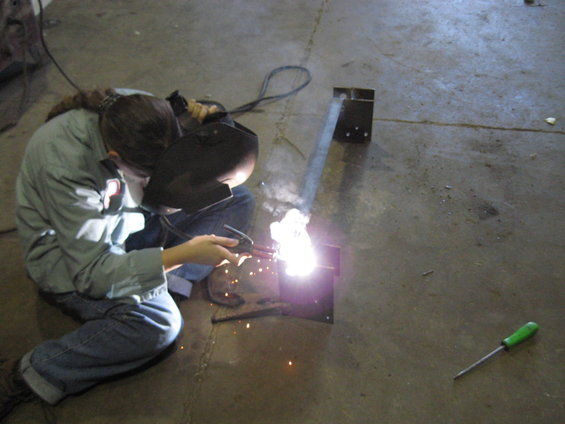 Person wearing a welding mask and jeans, sitting on the floor and welding a metal piece. Sparks fly from the weld