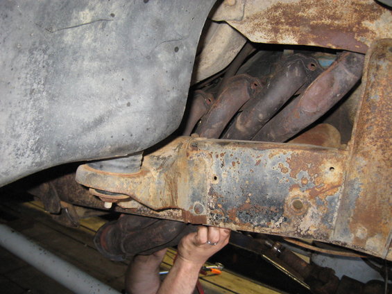 Close-up of rusty vehicle frame with exhaust manifold pipes visible. Hands are working on the frame