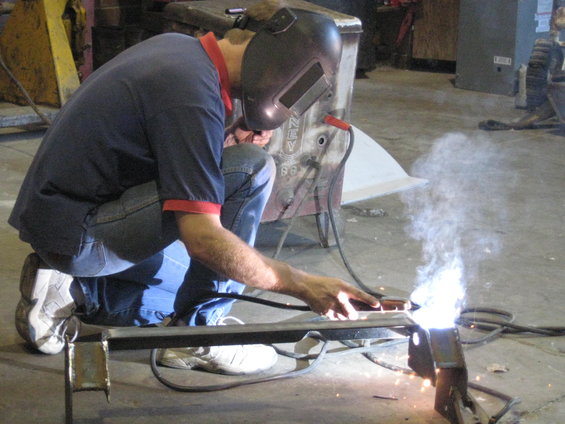 Welder in a blue shirt and jeans crouches, welding metal with sparks flying