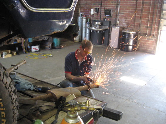 Man in garage welding exhaust pipe, sparks flying. Blue vehicle part above