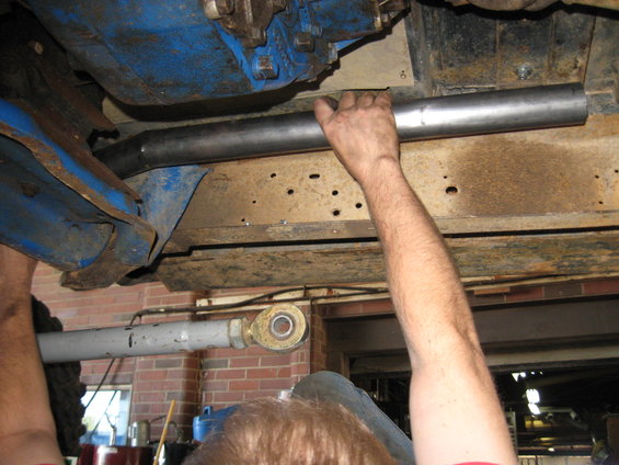 mechanic's hands installing a metal pipe under a vehicle's frame, showing blue painted components and rusty metal