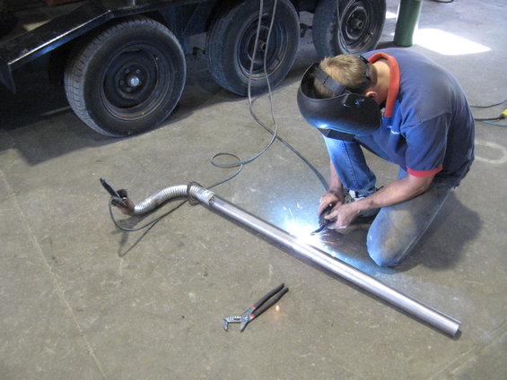 Guy welding a metal pipe with a flexible section, wearing a welding mask. A trailer with two wheels is in the background