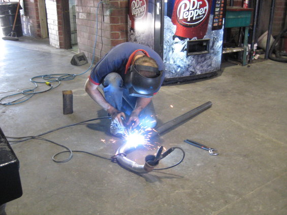 Welder sparks fly as exhaust pipe is welded. Dr Pepper vending machine in background