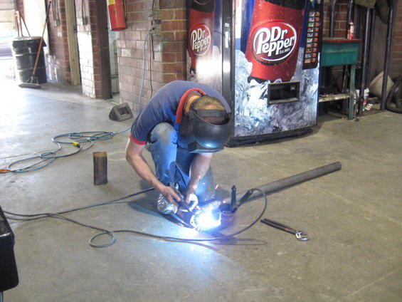 Person in welding mask working on metal pipe in garage with Dr Pepper vending machines in background