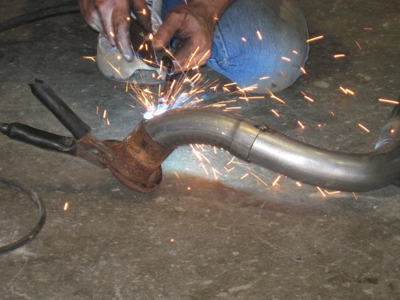 Exhaust pipe being welded with sparks flying. A person in blue jeans and a white shirt is welding