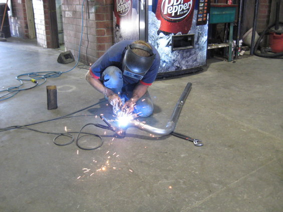 Man in welding mask working on a bent metal pipe on concrete floor, Dr Pepper vending machine in background