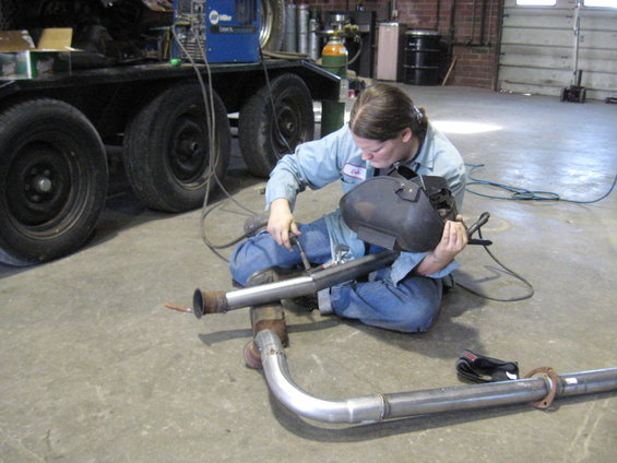 Person in blue work clothes and welding mask welding an exhaust pipe on the floor. A trailer with tires is in the background