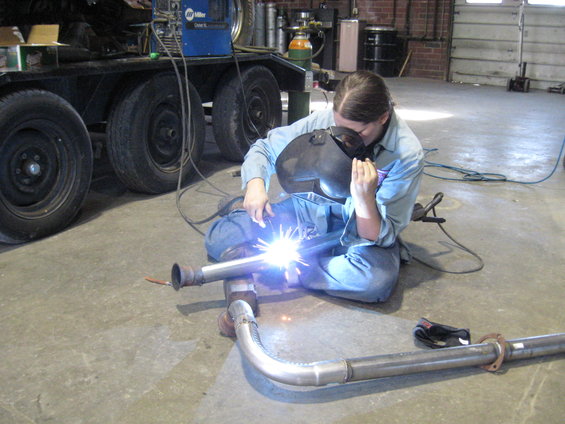 Person in welding mask working on exhaust pipe on concrete floor, trailer with tires in background
