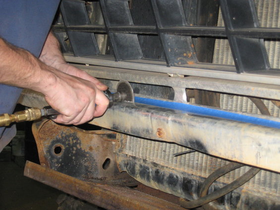 Hands using a cutting tool on a vehicle's front bumper, with the grille and radiator visible behind it