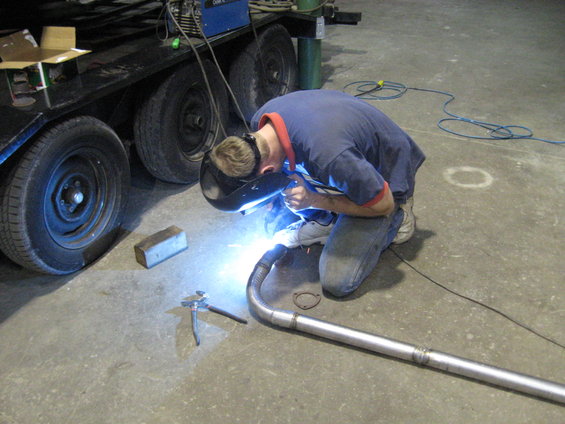 Man in welding mask working on exhaust pipe next to a trailer with multiple wheels