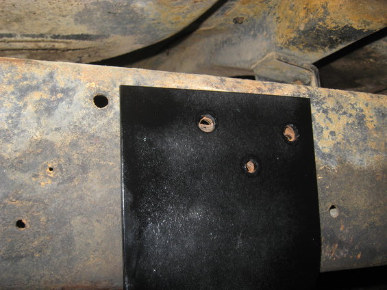 Black metal plate bolted to rusty vehicle frame with three visible holes