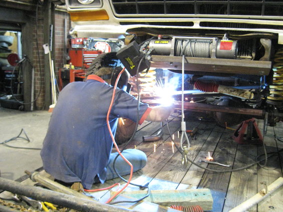 Guy welding on the frame of a vehicle, possibly a truck or SUV, in a garage. A winch is mounted to the front