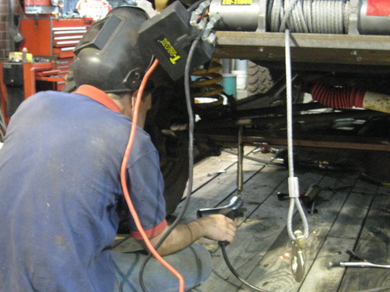 Person in welding mask working on vehicle undercarriage. A winch is visible above