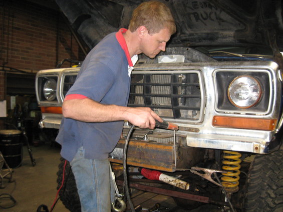 Guy welding a metal plate onto the front bumper of a large truck. Hood is up, showing engine bay