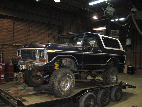 Dark blue 1978 Ford Bronco with white top, lifted with large tires, on a triple-axle trailer in a workshop