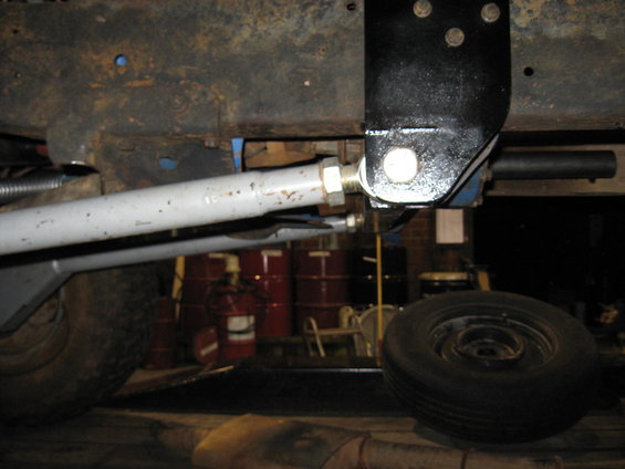 Underside of a vehicle with suspension components and a spare tire visible. Some red barrels are in the background