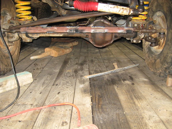 close-up of a vehicle's front axle, suspension, and exhaust system on a wooden floor