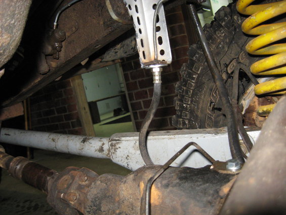 Close-up of a vehicle's rear axle, suspension, and brake line. A yellow coil spring is visible