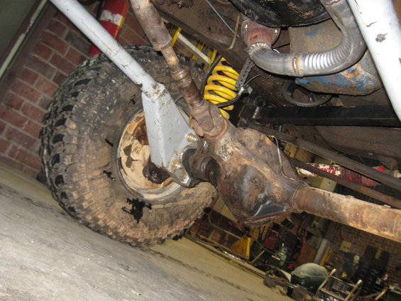 Undercarriage of a vehicle showing a large tire, rusty differential, and yellow coil spring