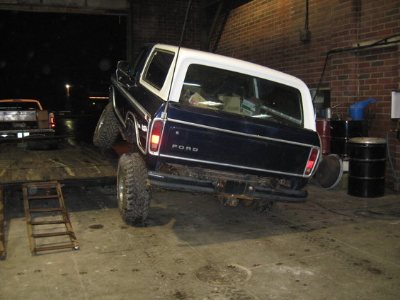 Dark blue Ford Bronco with white top, rear view, being loaded onto a trailer. Another truck is visible in the background