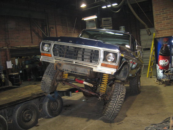 Blue Ford Bronco on a trailer, front end lifted. It's in a garage with a Dr. Pepper sign