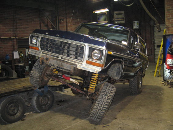 Blue 1978 Ford Bronco on a trailer, front-left wheel off the ground, showing its suspension