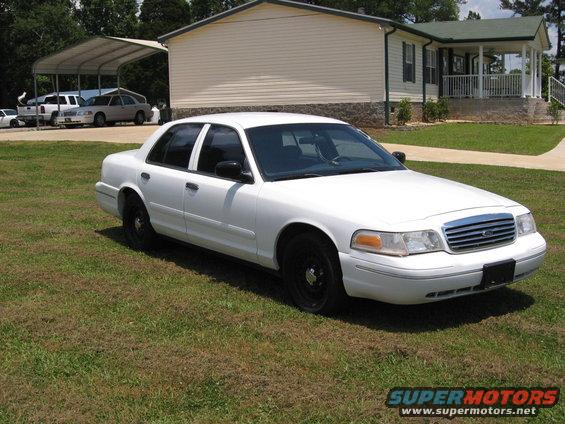 White sedan parked on grass, front 3/4 view. Black wheels, black side mirrors. House and carport in background