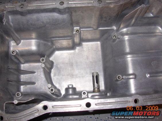 Close-up of an aluminum oil pan with mounting holes and internal baffles. A small metal tube protrudes from the bottom