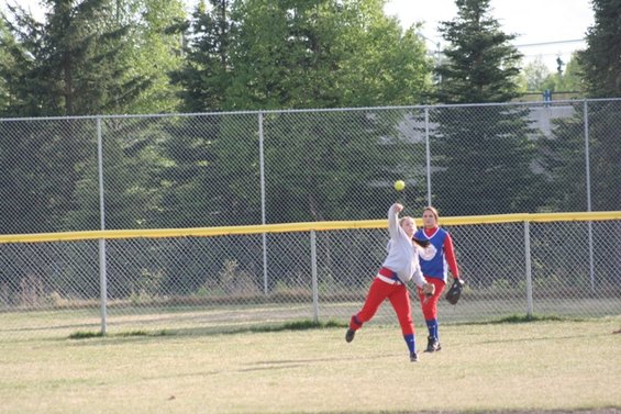 Two girls playing softball on a field with a chain-link fence and trees in the background