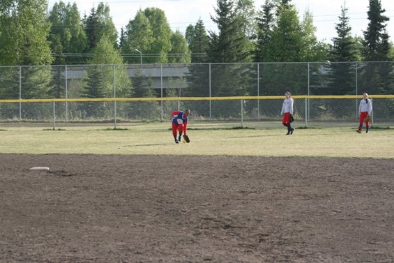 Players on a softball field, one in the foreground bending down, two others in the background