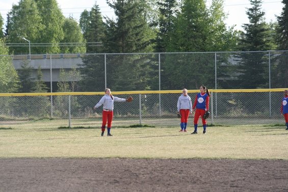 Three softball players in red pants and gray or blue tops stand on a grassy field behind a chain-link fence